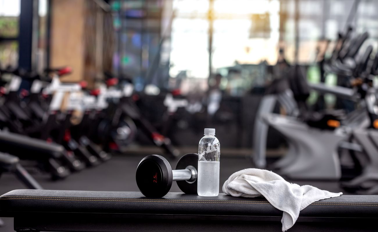 Eine Hantel, eine Wasserflasche und ein Handtuch liegen auf einer Bank in einem Fitnessstudio mit Trainingsgeräten im Hintergrund.