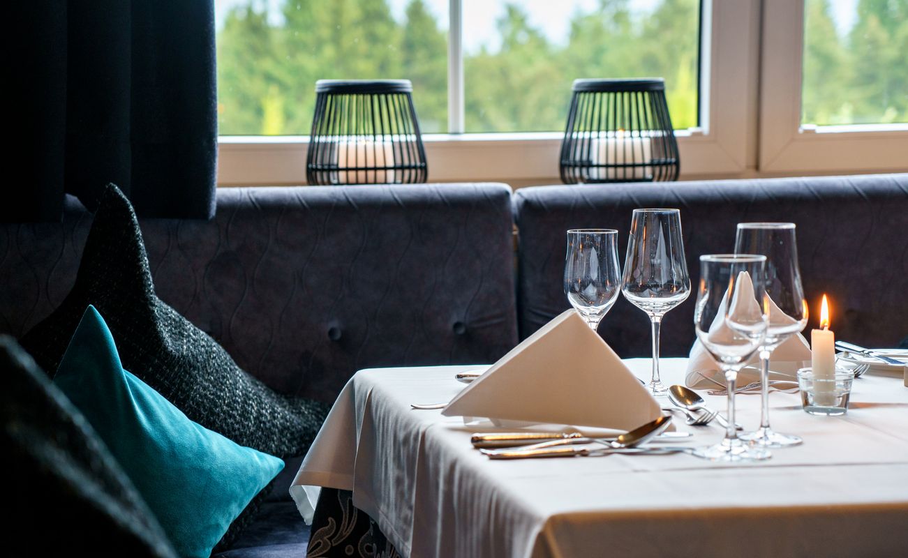 Elegant gedeckter Tisch in einem Restaurant mit Gläsern, Besteck und Servietten. Im Hintergrund sind Fenster und dekorative Lampen sichtbar.
