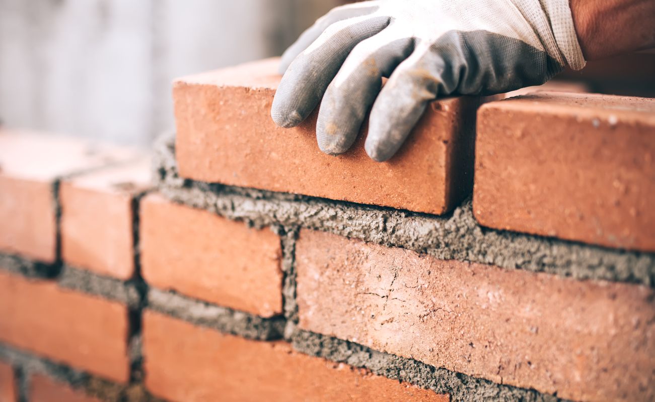 Eine Hand mit einem Handschuh platziert einen Ziegelstein auf einer Mauer aus Ziegeln und Mörtel.